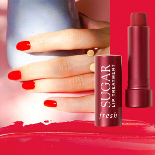 A hand with red nail polish next to an opened lip treatment case with a smearing of red lip treatment below.