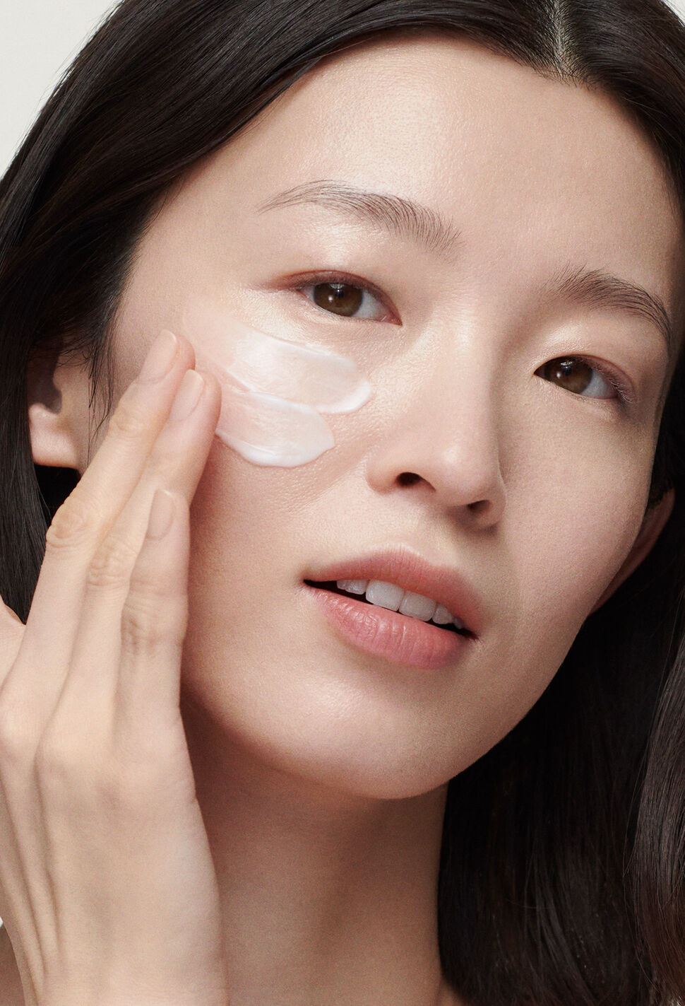 Woman applying white moisturizer to her cheekbone against grey background