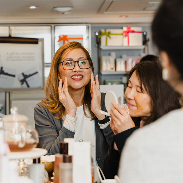 A women in glasses and her friend, who is holding a white compact mirror, try on Fresh products as they talk to someone who works in the shop